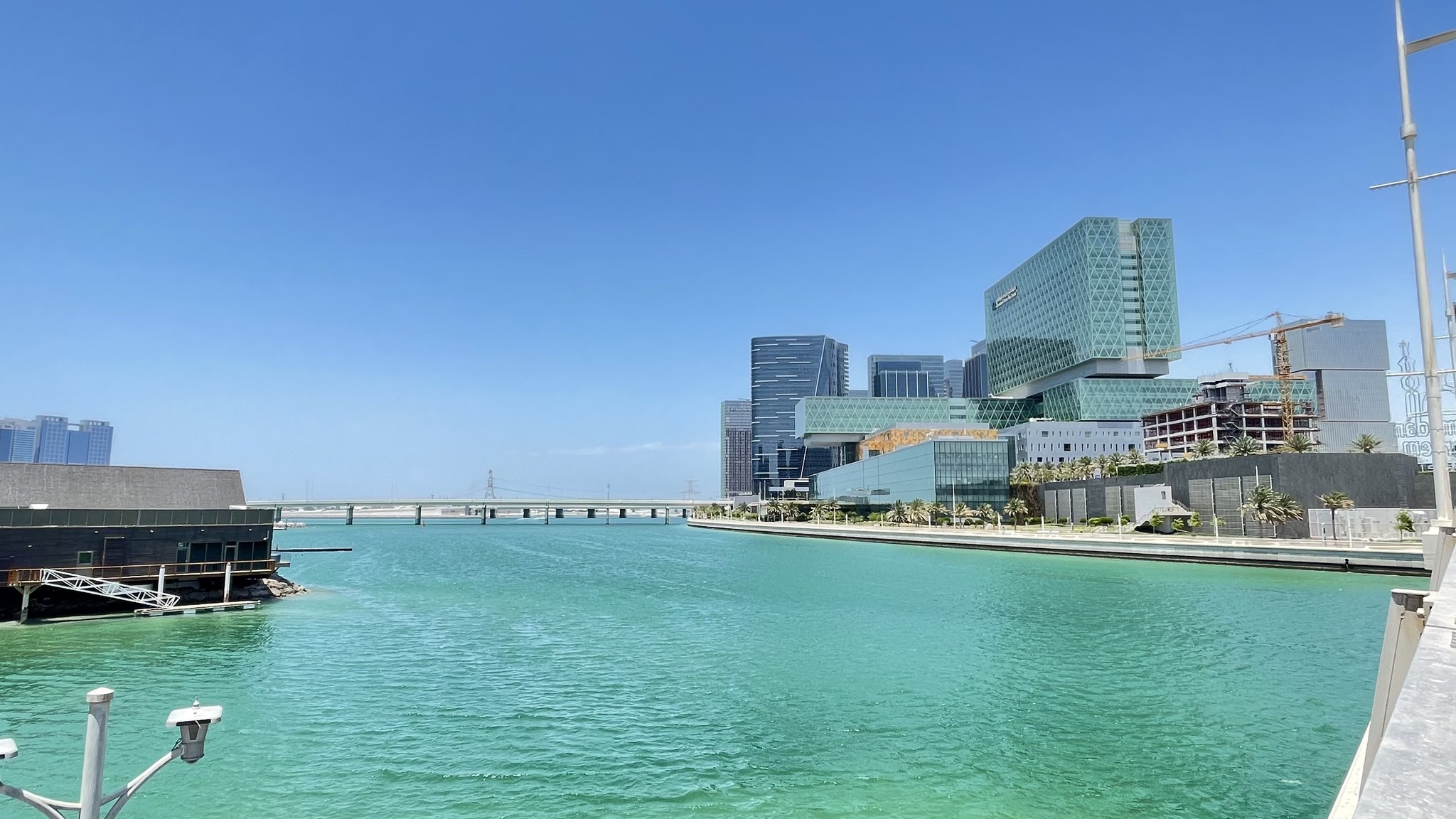 Office towers and a waterfront promenade on Al Maryah Island in Abu Dhabi, beside a turquoise channel under a clear sky.