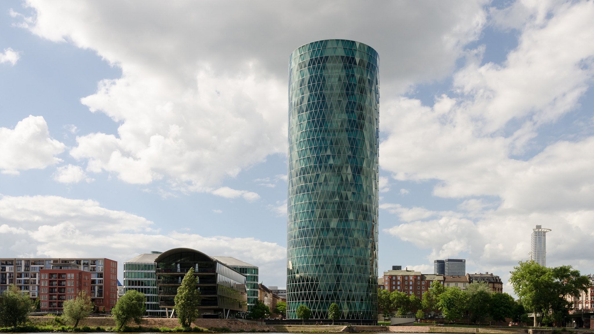 The cylindrical glass Westhafen Tower in Frankfurt am Main, with lower buildings and trees in front and a cloudy sky.