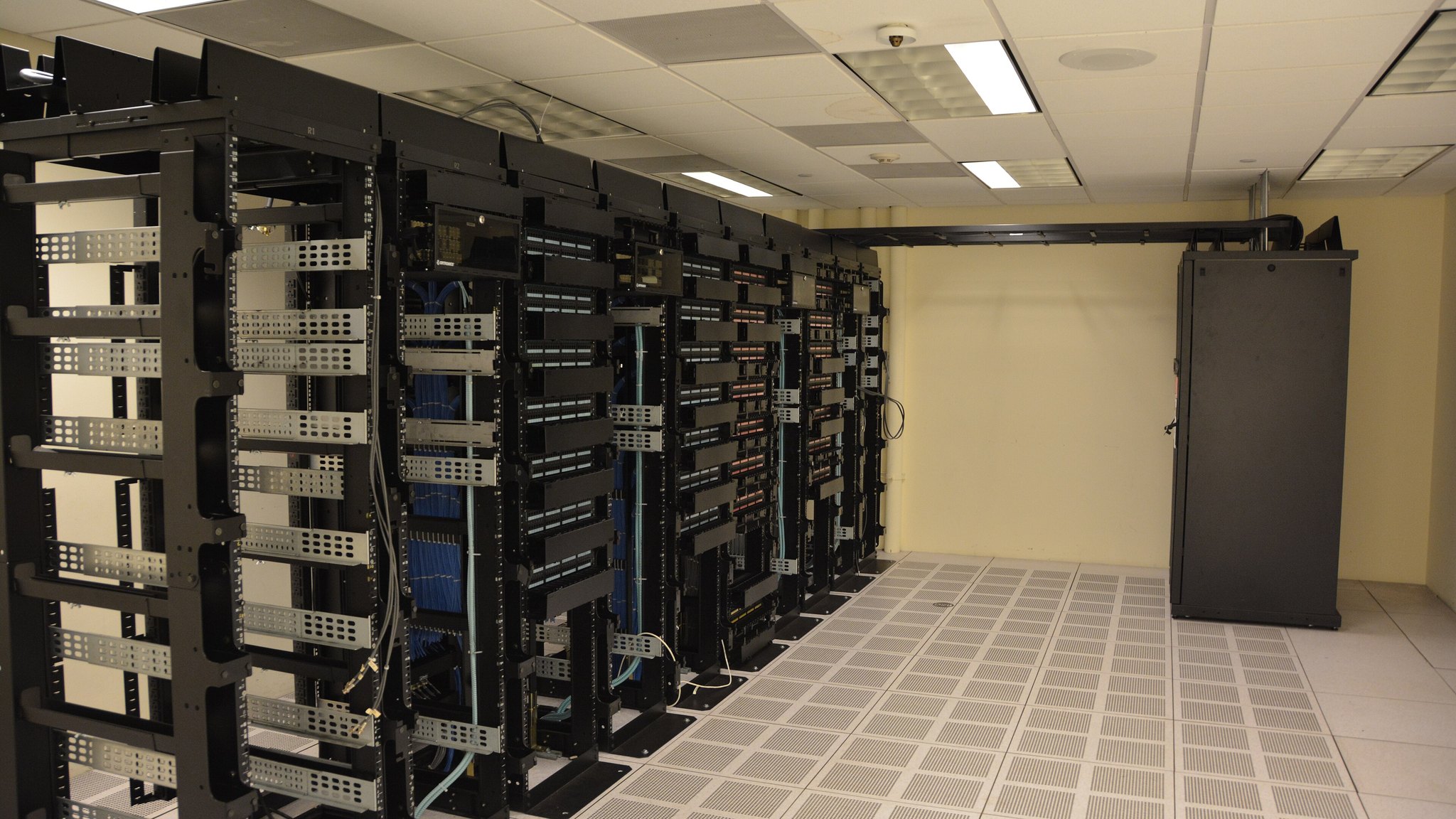 Rows of black server racks with cabling in a data centre room with a tiled floor.