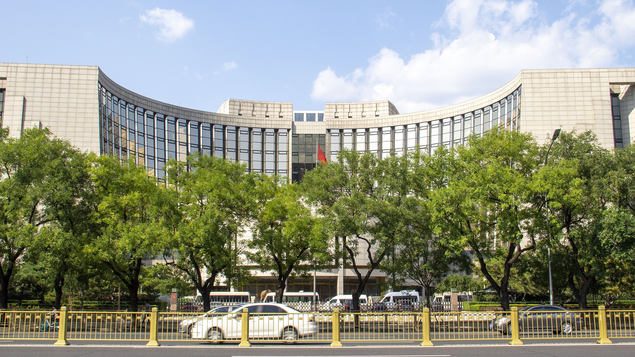 The curved headquarters building of the People's Bank of China in Beijing, behind trees along the street.